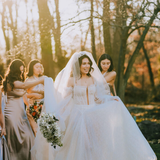 Bride walks through garden as bridal party helps carry the back of her gown.