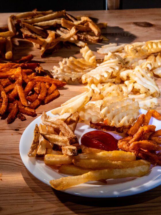 French fries, from sweet potatoes to potato wedges, displayed on wooden counter top and white plate.