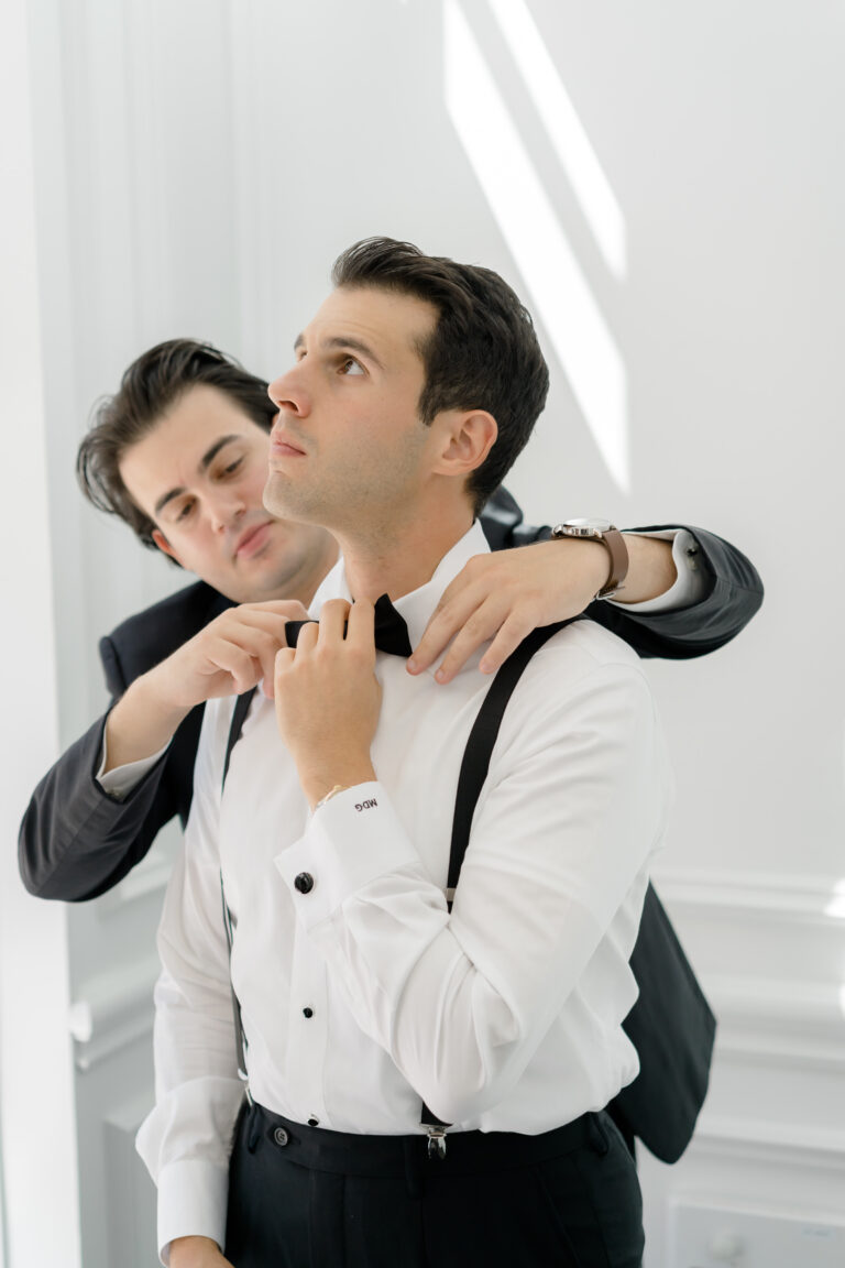 Groomsmen helps groom straighten bow tie as he gets ready.