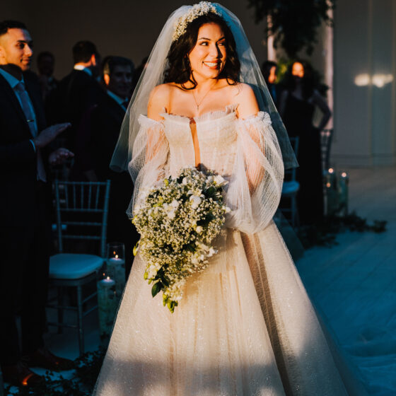 Bride smiles as she walks down sunset filled aisle during wedding ceremony.
