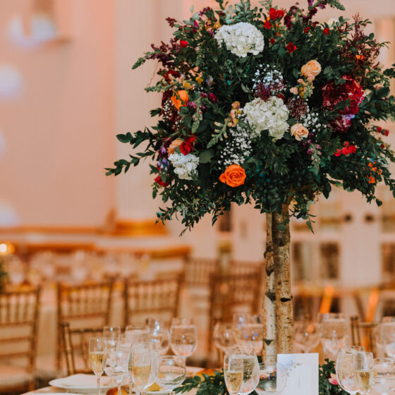 Reception table decorated with a greenery centerpiece filled with white hydrangeas, orange roses, and red accents.