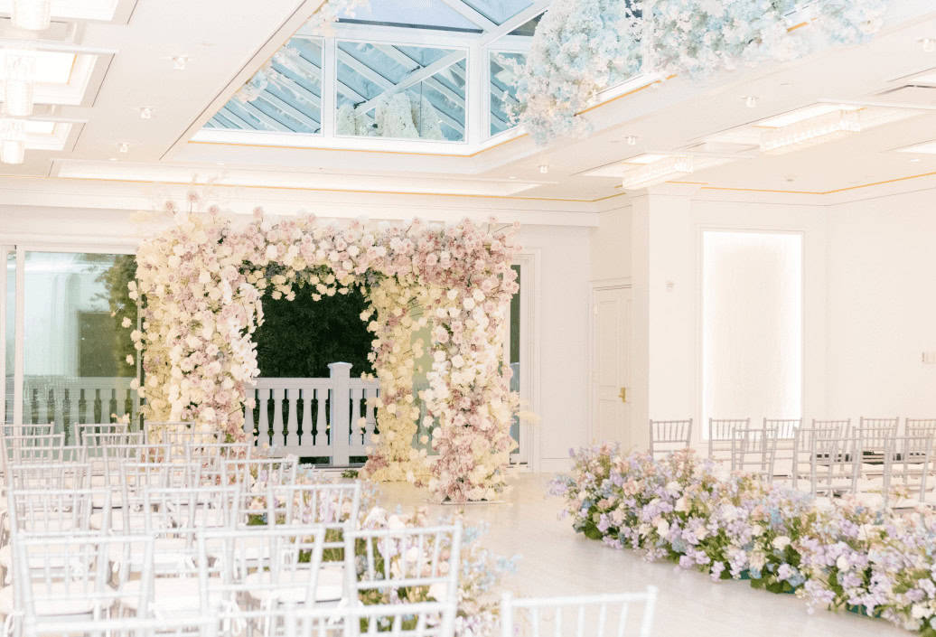 Spring flower arch in the Atrium filled with light pink and white roses. Aisle lined with pastel purple and blue flowers.