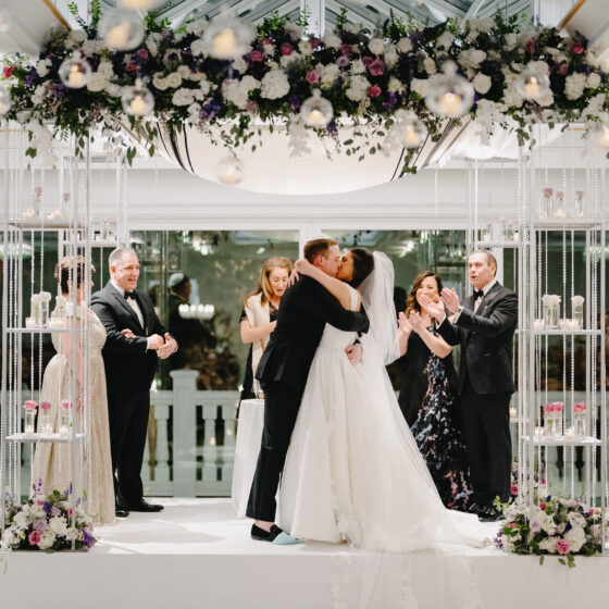 Bride and groom kiss underneath floral chuppah as just married couple.