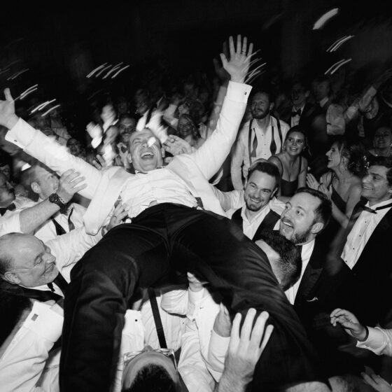 Black and white photo of groom lifted above crowd as he cheers with excitement.