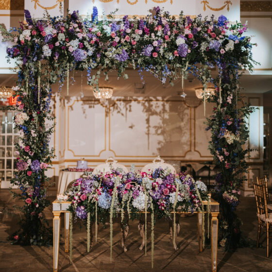 Sweetheart table elaborately decorated with a flowers and a flower arch filled with purple, pink, white, light blue flowers and greenery.