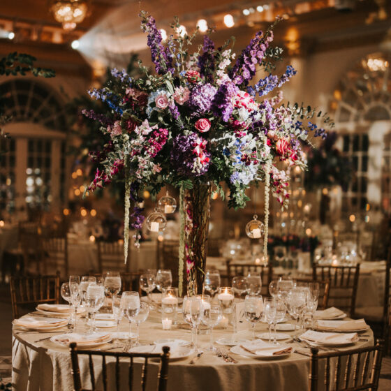 Wedding reception table is decorated with delicate tableware and a tall, colorful flower centerpiece with hanging tea lights.