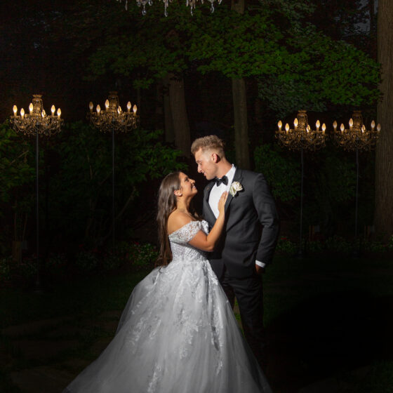 Bride and groom pose for wedding portrait underneath chandelier at night in Crystal Plaza's garden.