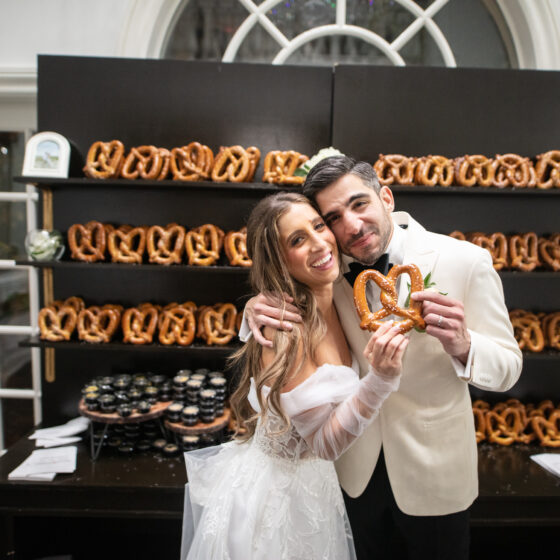 Bride and groom hold a pretzel in front of a gourmet pretzel wall.