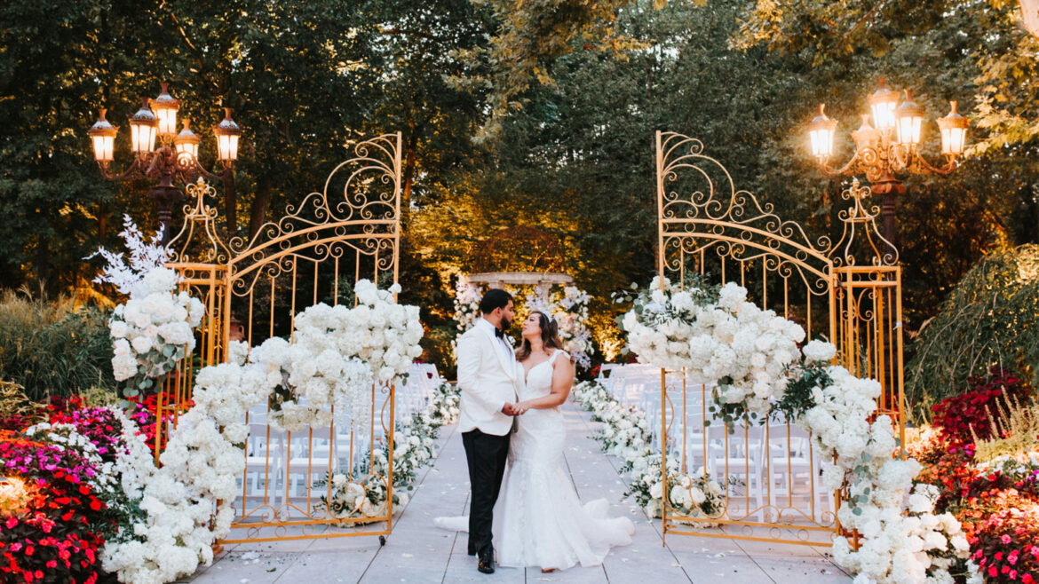 Bride and groom pose for photo in front of elaborately decorated gold arches with white flower in Crystal Plaza's garden.