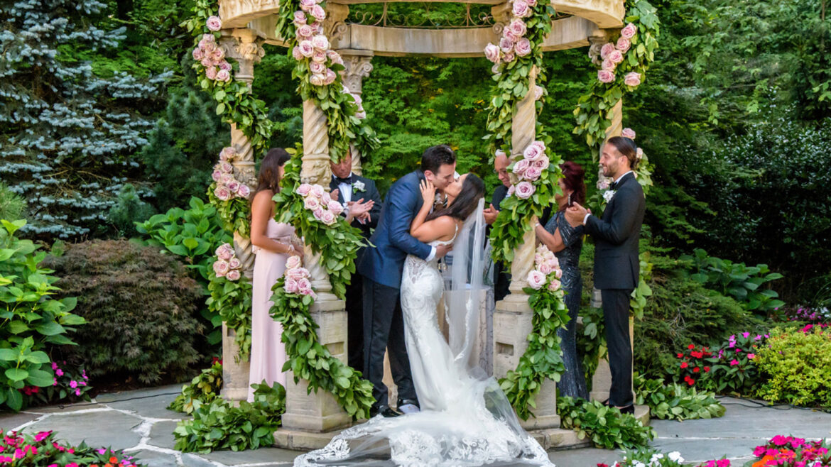 Bride and groom kiss under gazebo decorated with pink roses and greenery.