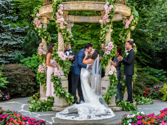 Bride and groom kiss under gazebo decorated with pink roses and greenery.