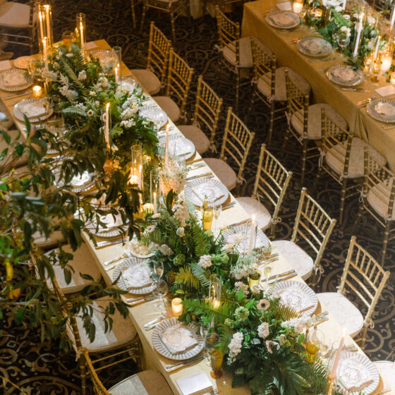 Overview shot of a long table, decorated with table runner filled with greenery, candles, and elegant tableware.