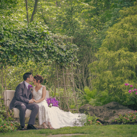 Groom kisses bride as they sit on a swinging bench in Crystal Plaza's gardens.