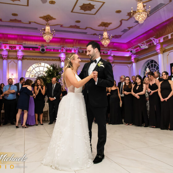 Bride and groom smile during first dance as guests gather round on the dance floor.