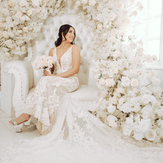 Bride sits in gorgeous white chair decorated with lush white roses.