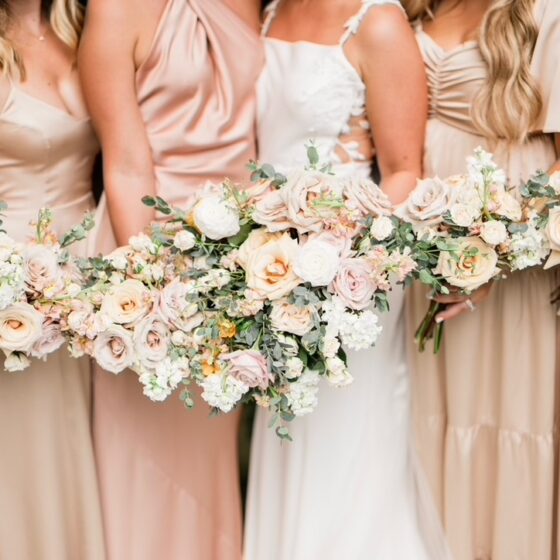 Bride and bridesmaids hold flower bouquets, filled with light pink, orange, and white roses.